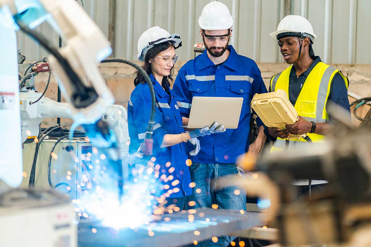 Three engineers in safety gear using a laptop to monitor an automated robotic welding process in a smart factory, illustrating how Private 5G and IoT enhance efficiency and connectivity in advanced manufacturing.
